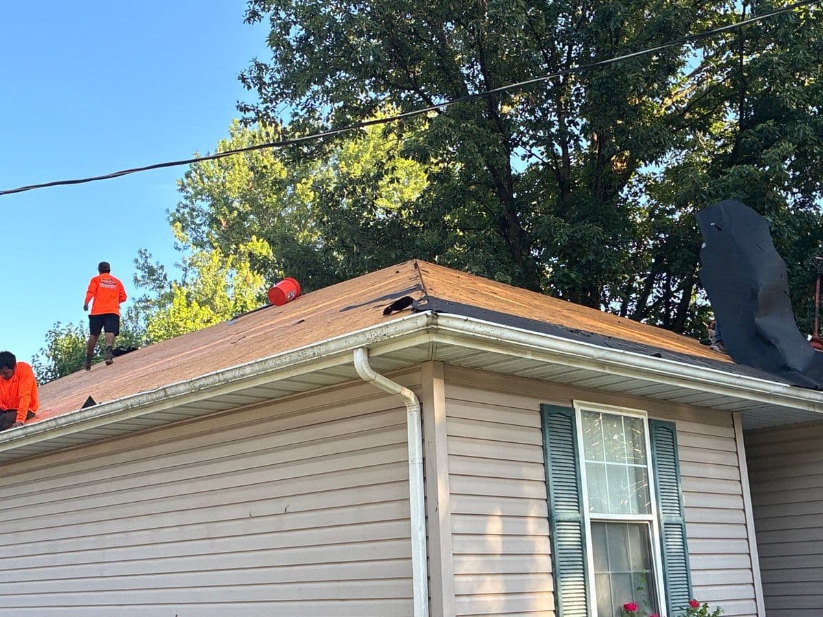 Two crew in orange shirts working a stripped roof deck under a clear blue sky