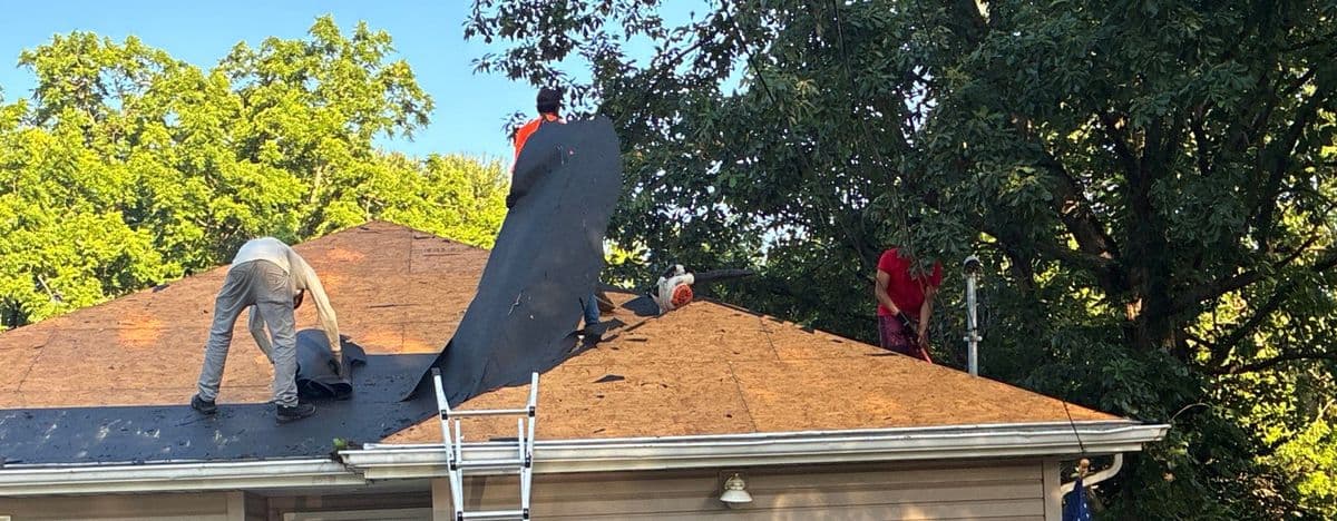 Crew pulling underlayment across the bare decking, tree line behind and a ladder at the eave