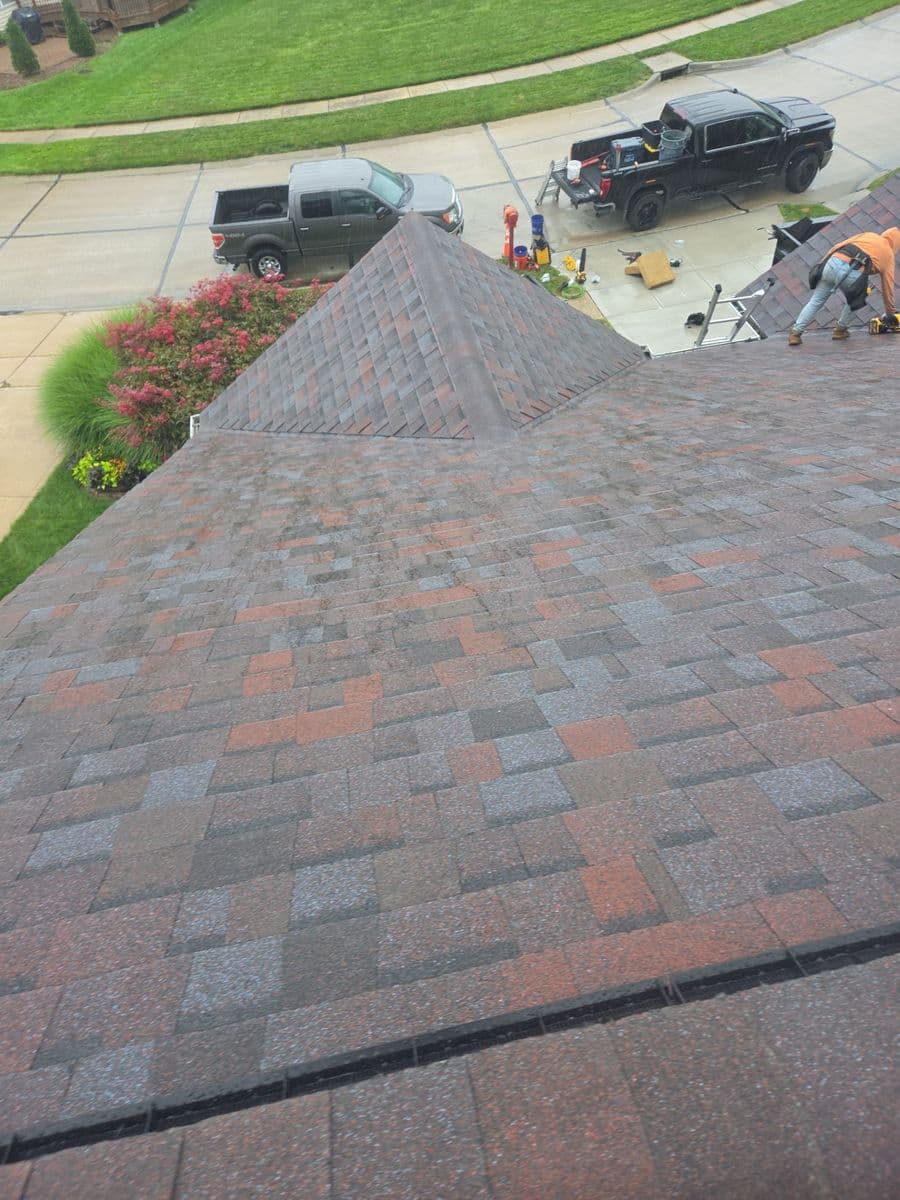 Finished red and grey blend shingle roof over a cul-de-sac, work trucks and a ladder in the driveway below